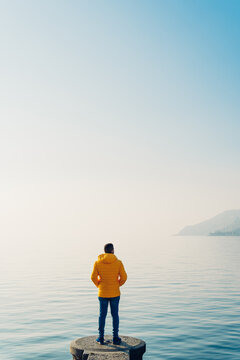 Anonymous guy on stone pier against peaceful sea waterBack view of unrecognizable male in outerwear standing on stone pier near tranquil sea under cloudless blue sky in Switzerland.