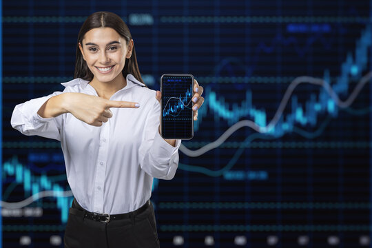 Happy younng european businesswoman pointing at mobile phone with glowing candlestick forex chart and index on dark grid background. Trade, stock and finance concept. Double exposure.