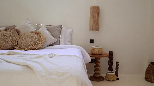 Room decor details in Boho villa bedroom with throw cushions, wooden nightstand, hanging lamp, straw woven basket, and totems on a floor - dolly left
