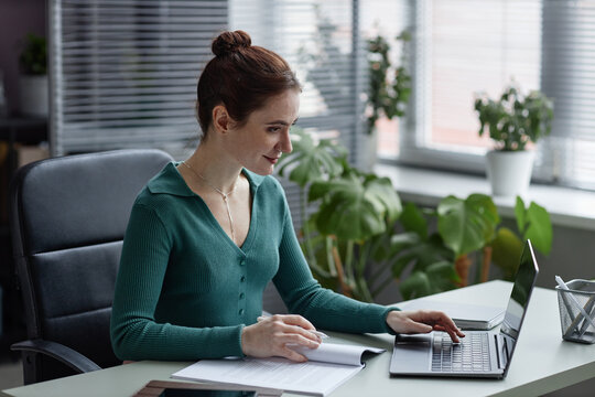 Side view portrait of smiling young woman working in cozy office decorated with green plants
