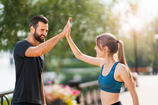 Image of happy smiling couple giving highfive, high five hands gesture, woman with man or bearded coach trainer, clapping palms, after successful training, outdoors. Fitness, sport, workout concept.