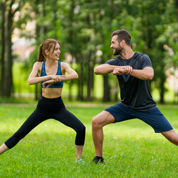 Square image - young smiling couple, woman training with man or bearded coach trainer, do squat fit exercise together, look at each other, outdoors. Fitness, sport, workout, healthy lifestyle concept.