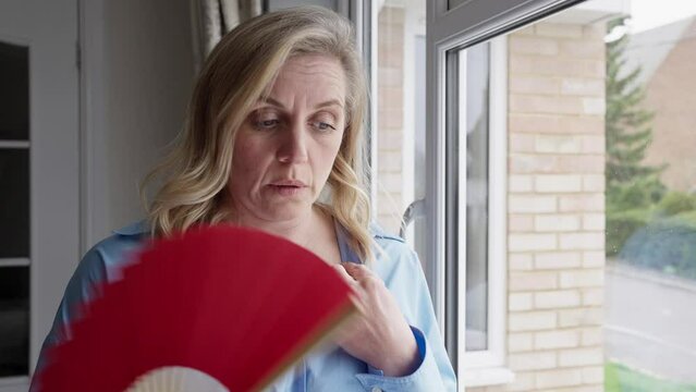 Menopausal Mature Woman Having Hot Flush At Home Cooling Herself With Handheld Paper Fan 
