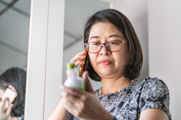 Asian woman is listening to a doctor advise medical use from her smartphone, her hand is holding a liquid medicine bottle, eyes looking at the bottle. Asian middle-aged woman wears eyeglasses.