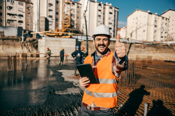 A constructor is standing on building foundation with tablet in his hands and giving thumbs up and smiling at the camera. The workers in background are concreting foundation.