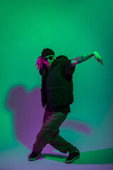 Fototapeta na wymiar Cool trendy professional hip hop dancer guy with a hat and sunglasses in fashion black clothes with a vest dances in motion in a creative color studio with green and pink light
