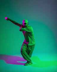 Stylish fashion dancer professional man with a cap in trendy outfit with sneakers dances in motion in a creative studio with green and pink light