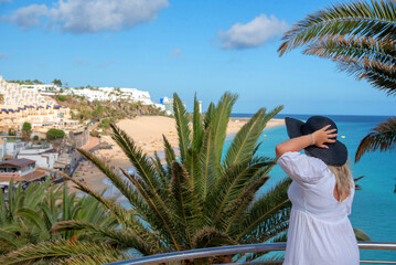 Mujer rubia con un vestido blanco sosteniendo un sombrero mientras mira la playa de arena blanca y...