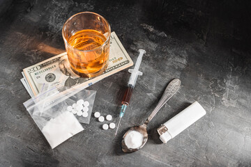 Alcohol drink in a glass, syringe with a dose of drugs, white pills in a transparent bag, narcotics powder in a spoon and US dollar cash on dark background. Concept of addiction and bad habits