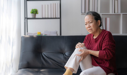 Asian elderly woman sitting with knee pain on sofa at home.