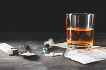 Alcohol drink in a glass, syringe with a dose of drugs, white pills in a transparent bag, narcotics powder in a spoon and US dollar cash on dark background. Concept of addiction and bad habits