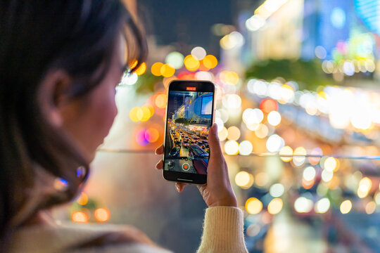 Young Asian woman using mobile phone taking picture of urban cityscape with street lights at night. Attractive girl enjoy and fun outdoor nightlife travel city with using portable device technology.