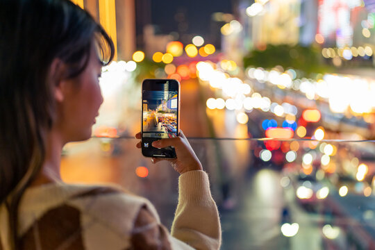 Young Asian woman using mobile phone taking picture of urban cityscape with street lights at night. Attractive girl enjoy and fun outdoor nightlife travel city with using portable device technology.