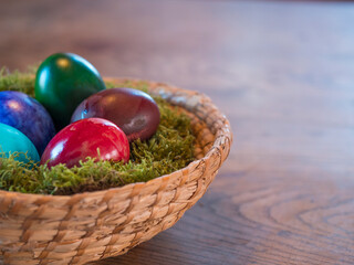 Various coloured decorated easter eggs in wicker basket nest with moss and colored Easter eggs on beige wooden bokeh background. For Happy Easter holiday card. Selective focus, copy space