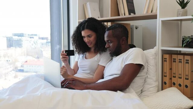 Loving multiethnic family spending morning in bed with laptop while using credit card for internet banking at home. Romantic partners making online payment operations in available application.