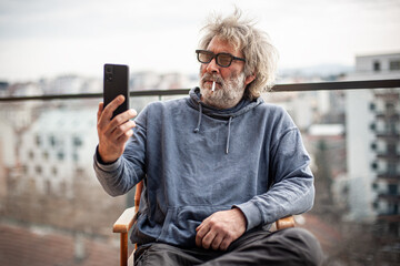 Handsome senior man with grey hair and eyeglasses sitting on terrace, using cigarette and smartphone, communicating, having fun and enjoying