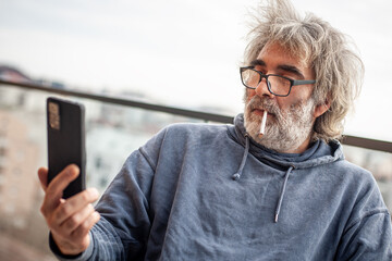 Handsome senior man with grey hair and eyeglasses sitting on terrace, using cigarette and smartphone, communicating, having fun and enjoying
