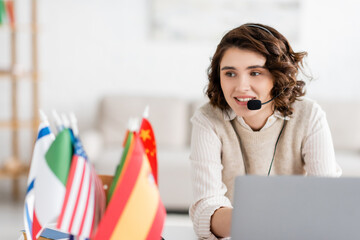 young language teacher in headset smiling during online lesson near laptop and international flags...