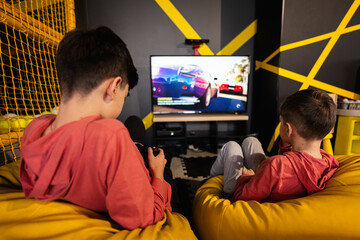 Two brothers playing race video game console, sitting on yellow pouf in kids play center.