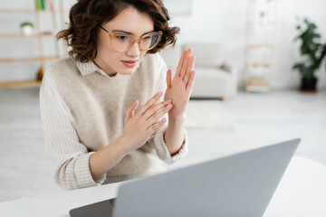 young teacher in glasses showing two handed sign language gesture while looking at laptop. © LIGHTFIELD STUDIOS
