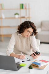curly student in glasses holding smartphone while taking notes near laptop at home.