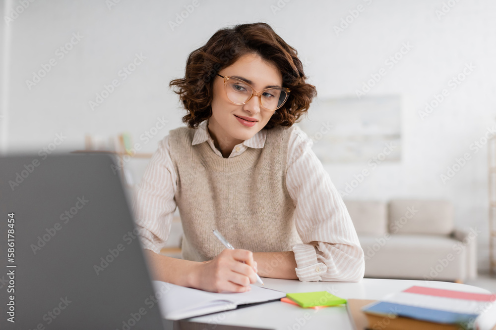Wall mural smiling woman in glasses taking notes while learning foreign language and watching online lesson on 