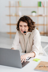 young and happy language teacher in headset typing on laptop while having online lesson at home.