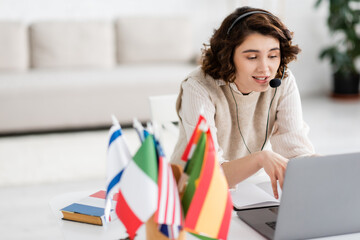positive language teacher in headset talking during online lesson on laptop near international flags at home.