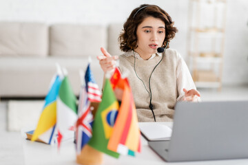 language teacher in headset pointing with fingers while talking during video chat on laptop near blurred flags at home. © LIGHTFIELD STUDIOS