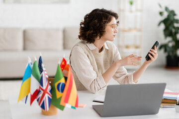 young language teacher in glasses holding smartphone near laptop and international flags on blurred foreground.