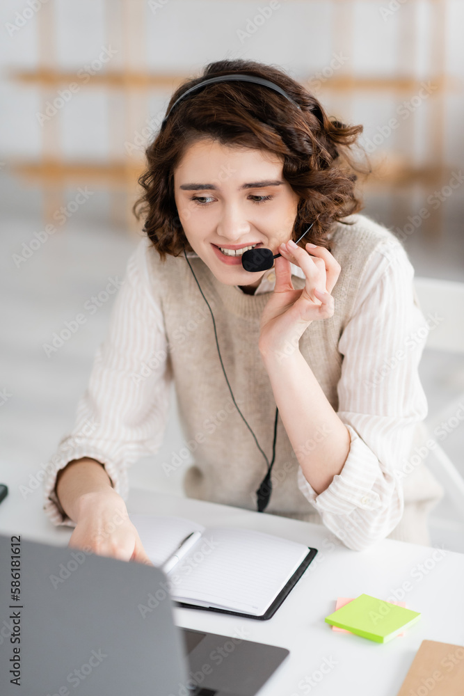 Wall mural smiling language teacher in headset talking near laptop and blank notebook while working at home.