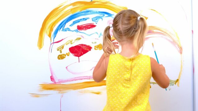 child enthusiastically draws with artistic brush confident strokes paint on white wall home, blonde preschool girl in yellow dress paints rainbow, flowers, childish naive drawing, happiness childhood