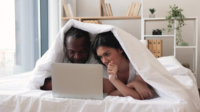 Young romantic partners resting on bed under white sheets while surfing internet via portable computer on sunny day at home. Multicultural married couple enjoying comfortable lifestyle at leisure.
