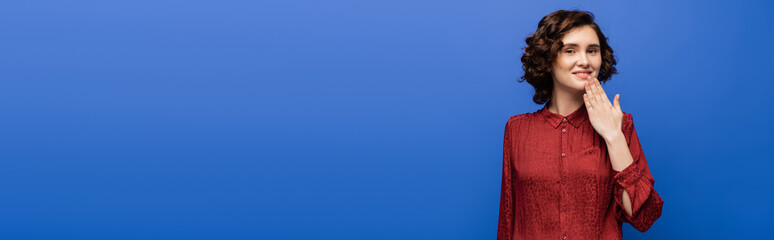 cheerful woman in red blouse telling thank you on sign language isolated on blue, banner.