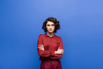 pretty language teacher with wavy hair looking at camera while standing with crossed arms isolated on blue.