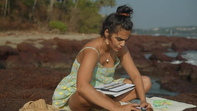 Woman in dress sitting on rocks by the sea preparing painting material.