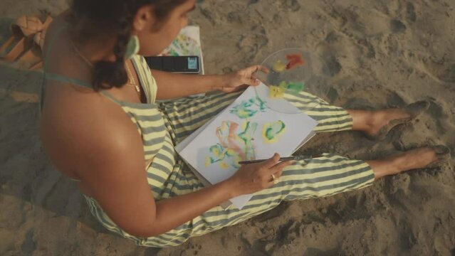 Young woman in striped top and pants sitting on the sand at the beach, painting with paint. Slow motion.
