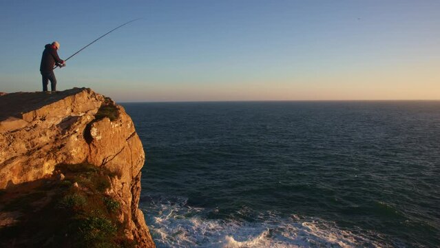 A man stands on a cliff by the sea and fishing at sunset