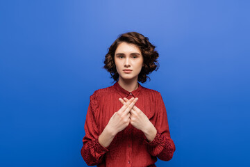 young brunette woman looking at camera while using sign language isolated on blue.