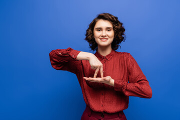 brunette woman smiling and showing gesture meaning stand on sign language isolated on blue.