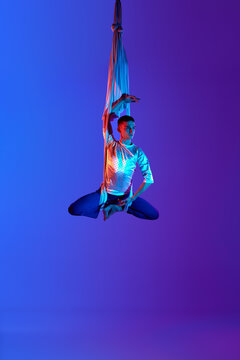 Male acrobat, professional, artistic aerial gymnast hanging upside down on aerial silk against gradient blue purple background in neon light. Concept of art, sportive lifestyle, hobby, action, motion