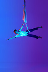 Acrobat, professional male aerial gymnast hanging upside down on aerial silk against gradient blue purple background in neon light. Concept of art, sportive lifestyle, hobby, action and motion, beauty