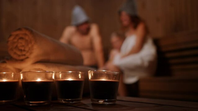 Close-up candles burning in Finn sauna with joyful Caucasian family having fun at background. Unrecognizable man and woman tickling girl laughing enjoying leisure with family indoors