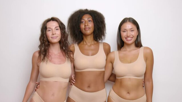 three young multiethnic women are standing in a studio against a white wall, dressed in beige underwear, they are joyfully rocking their hips from side to side
