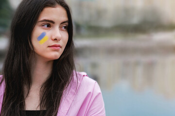 Thoughtful brunette teenage girl with ukrainian flag on face looking at distance at city park side...