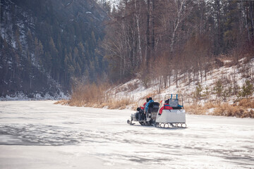 Winter walk with family on a snowmobile on a frozen river. A snowmobile with a trailer rolls children on the ice of the frozen Siberian Mana River in early spring.