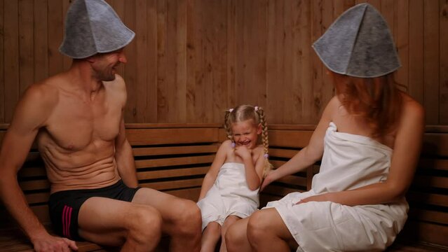 Loving father and mother kissing cheeks of smiling daughter sitting on wooden bench in Finn sauna. Happy carefree relaxed Caucasian family enjoying leisure in SPA indoors