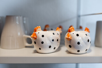 Two funny cute egg cups in the form of chickens stand decoratively on the kitchen shelf with cups in the background
