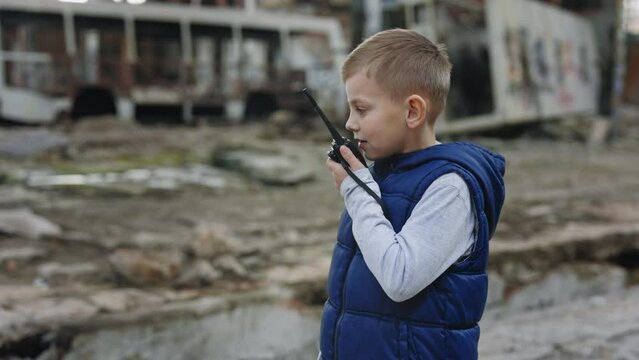 Boy Is Holding a Walkie-Talkie and Talking into It Standing in the Destroyed Plant. Child Gives Commands. The Game of Soldiers. War. The Little Commander