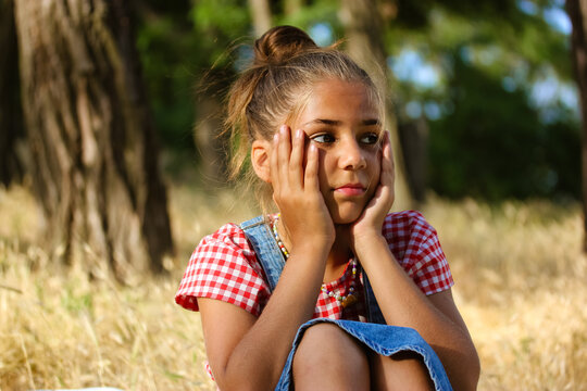 A beautiful little girl of school age 10-12 years old is sitting in a clearing in the woods in summer day. A child is bored on summer vacation. Problems in a family, child psychology, boring concept.